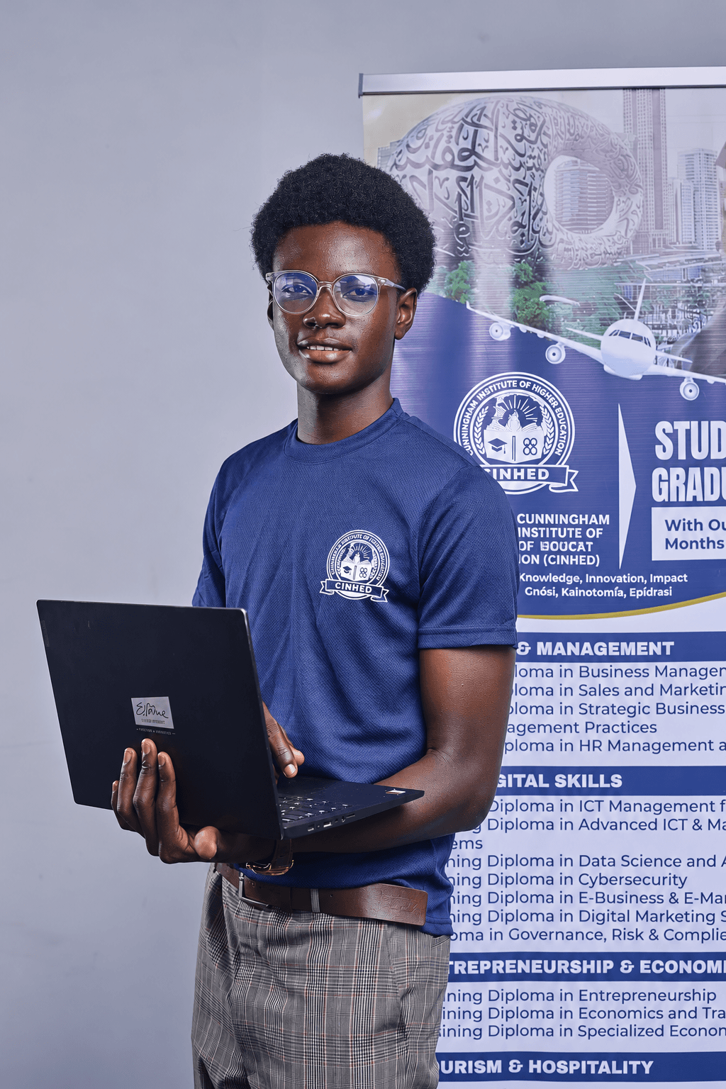 A CInHEd student holding a laptop in front of a programmes banner